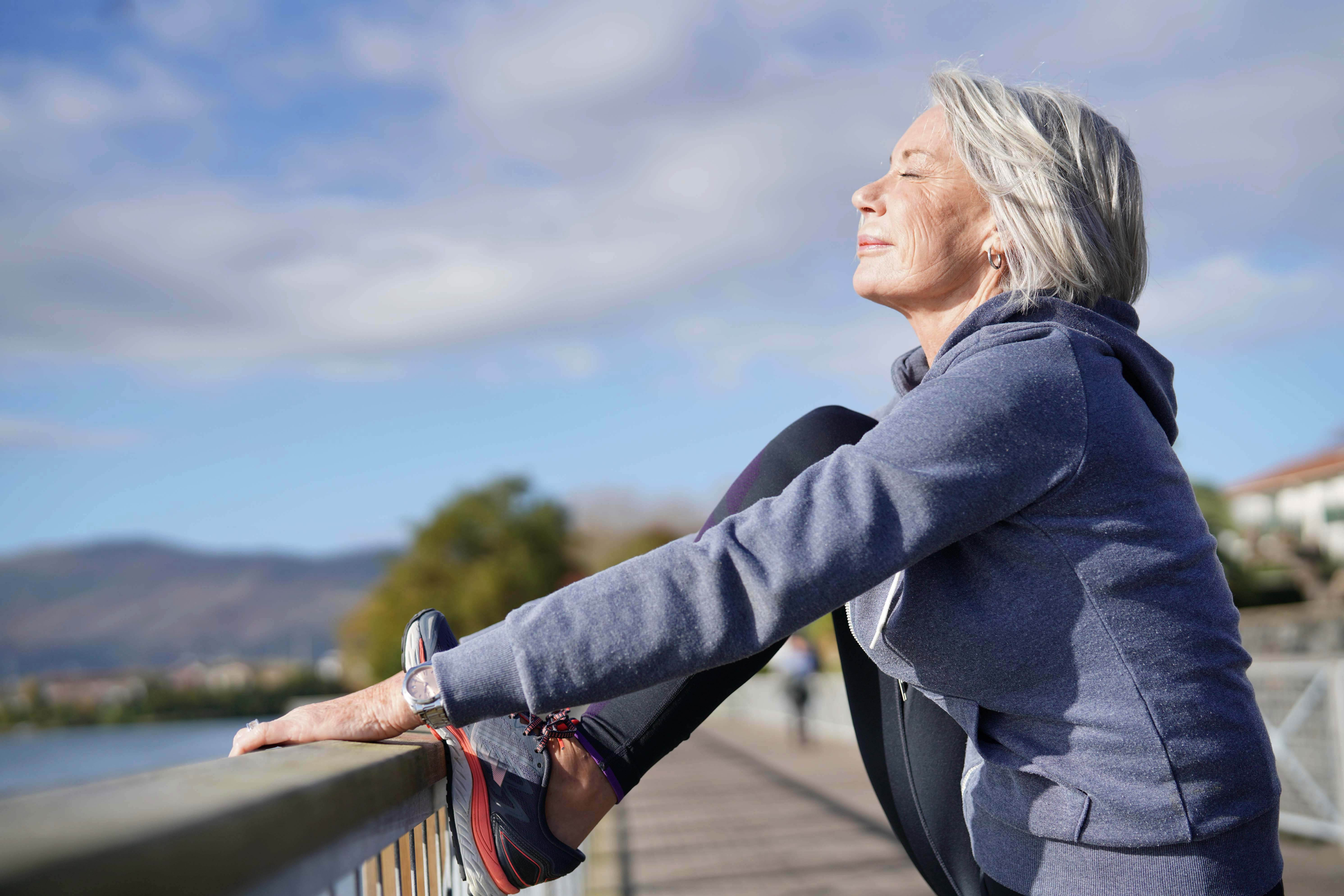 Older woman wearing a hoodie stretching her leg on a barrier near the water, with her face tilted up towards the sun and eyes closed Older woman wearing a hoodie stretching her leg on a barrier near the water, with her face tilted up towards the sun and eyes closed